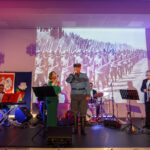 Grupa muzyków na scenie, w tle czarno-białe zdjęcie żołnierzy. Dominują kolory czerwony i biały z flagą Polski. Sala oświetlona fioletowym światłem.