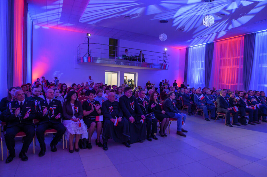 Na zdjęciu widać dużą salę wypełnioną ludźmi. Ściany są oświetlone na niebiesko i czerwono. Uczestnicy siedzą na krzesłach, niektórzy trzymają polskie flagi.