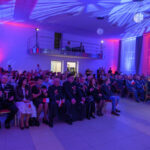 Na zdjęciu widać dużą salę wypełnioną ludźmi. Ściany są oświetlone na niebiesko i czerwono. Uczestnicy siedzą na krzesłach, niektórzy trzymają polskie flagi.