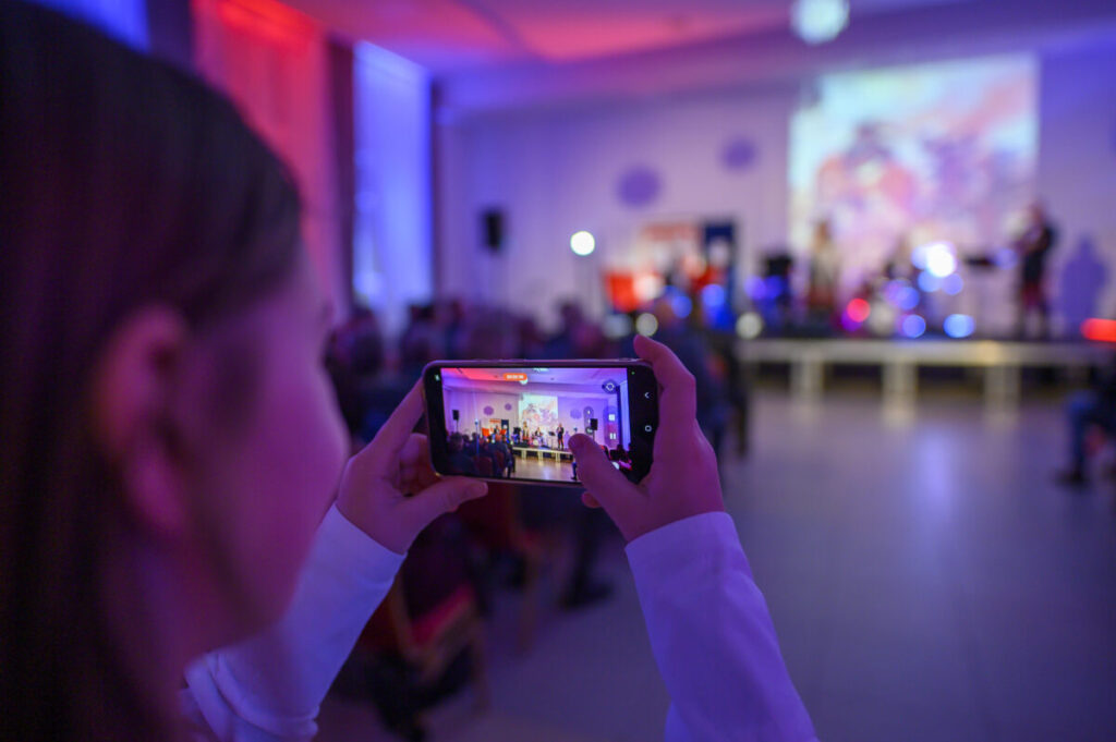 Zdjęcie przedstawia osobę robiącą zdjęcie sceny smartfonem. Kolory dominujące to niebieski i czerwony z rozmytym tłem. Na scenie widoczne sylwetki ludzi i projekcja świetlna.