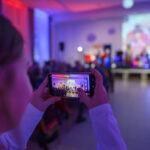 Zdjęcie przedstawia osobę robiącą zdjęcie sceny smartfonem. Kolory dominujące to niebieski i czerwony z rozmytym tłem. Na scenie widoczne sylwetki ludzi i projekcja świetlna.