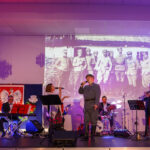 Na scenie występuje zespół muzyczny; tło fioletowe, flaga Polski obok. Projekcja starych fotografii żołnierzy w tle.