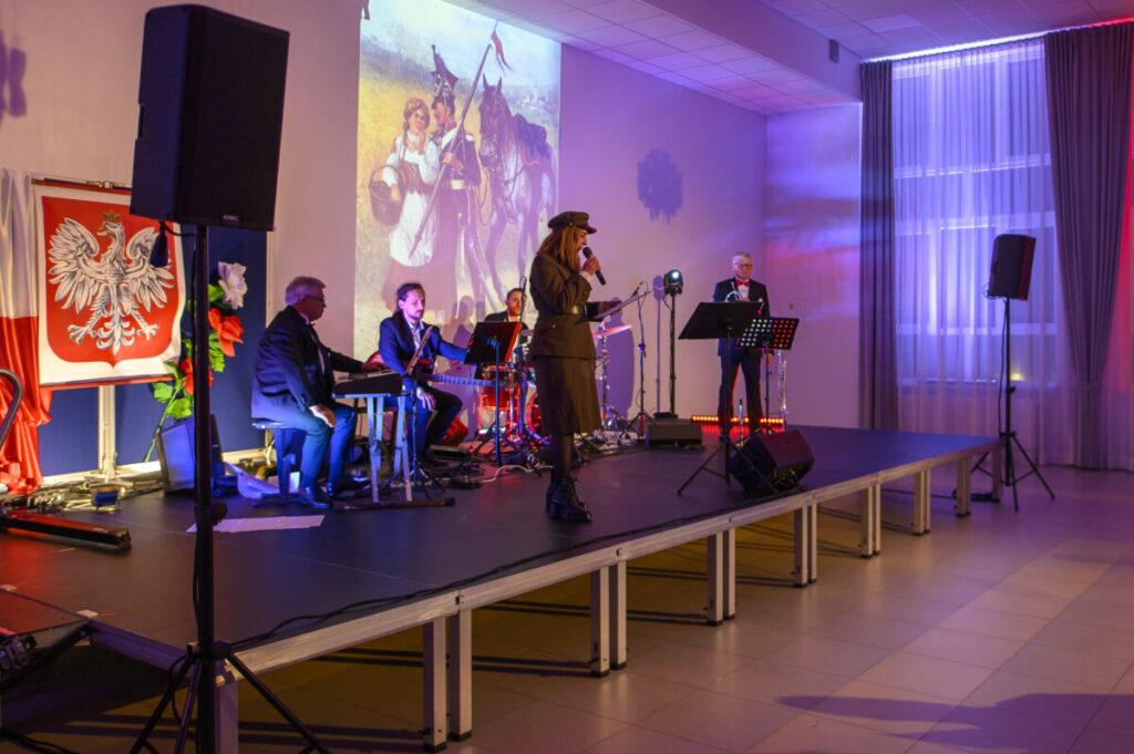 Grupa muzyków na scenie, z czerwonym i niebieskim oświetleniem. Tło stanowi obraz z końmi i postaciami historycznymi. Obok wisi biało-czerwona flaga z orłem.