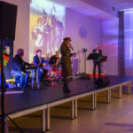 Grupa muzyków na scenie, z czerwonym i niebieskim oświetleniem. Tło stanowi obraz z końmi i postaciami historycznymi. Obok wisi biało-czerwona flaga z orłem.