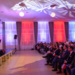 W sali koncertowej osoby siedzą na krzesłach z biało-czerwonymi flagami. Scena oświetlona jest fioletowo i czerwono. Na scenie stoją wykonawcy w eleganckich strojach.