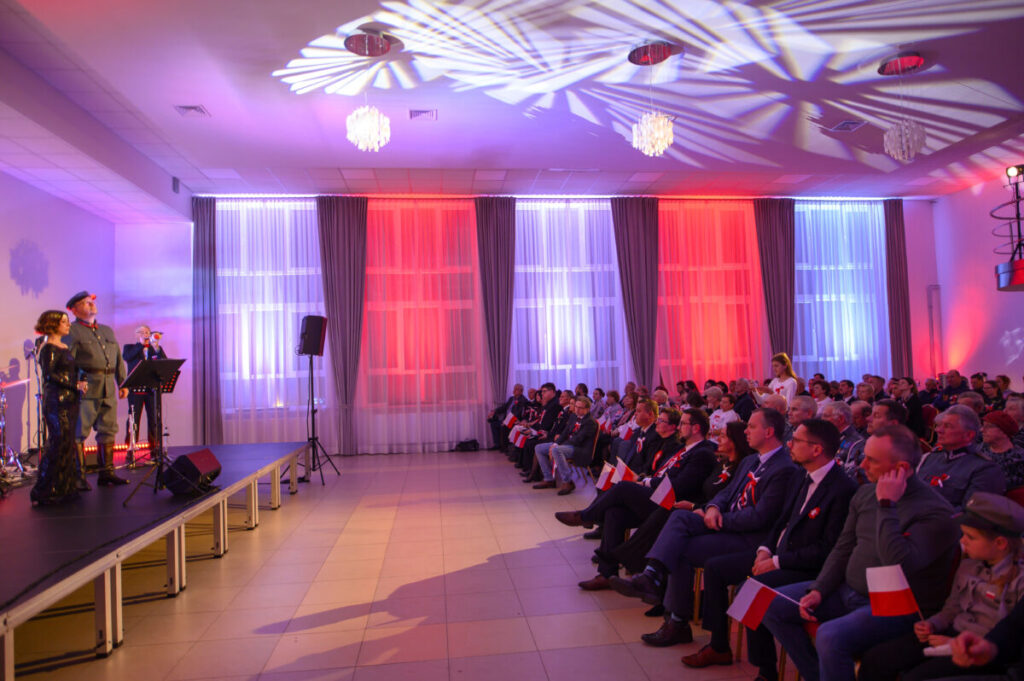 Koncert z wojskowymi i publicznością, sala w kolorach biało-czerwonych. Tło to oświetlone zasłony, osoby z polskimi flagami.