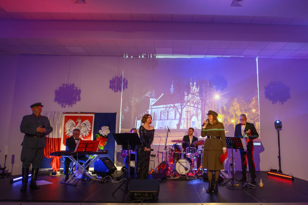 Na scenie zespół muzyczny, tło z polskim godłem i zdjęciem kościoła. Kolorowe oświetlenie sceniczne w tonach fioletu i czerwieni. Uczestnicy w eleganckich strojach.