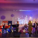 Na scenie zespół muzyczny, tło z polskim godłem i zdjęciem kościoła. Kolorowe oświetlenie sceniczne w tonach fioletu i czerwieni. Uczestnicy w eleganckich strojach.