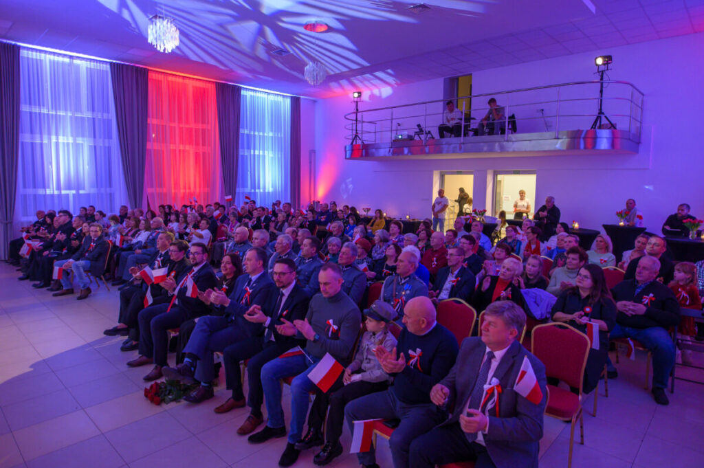 Na zdjęciu widzimy salę z tłumem ludzi trzymających małe flagi Polski. Oświetlenie w kolorach: niebieskim, czerwonym i białym. Tło to sceniczne dekoracje i balustrada na drugim planie.