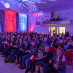 Na zdjęciu widzimy salę z tłumem ludzi trzymających małe flagi Polski. Oświetlenie w kolorach: niebieskim, czerwonym i białym. Tło to sceniczne dekoracje i balustrada na drugim planie.