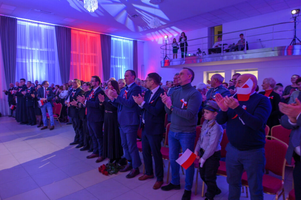 Grupa ludzi stoi i bije brawo; sala w biało-czerwonych światłach. Wiele osób trzyma polskie flagi. W tle balkon z obserwatorami.