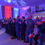 Grupa ludzi stoi i bije brawo; sala w biało-czerwonych światłach. Wiele osób trzyma polskie flagi. W tle balkon z obserwatorami.