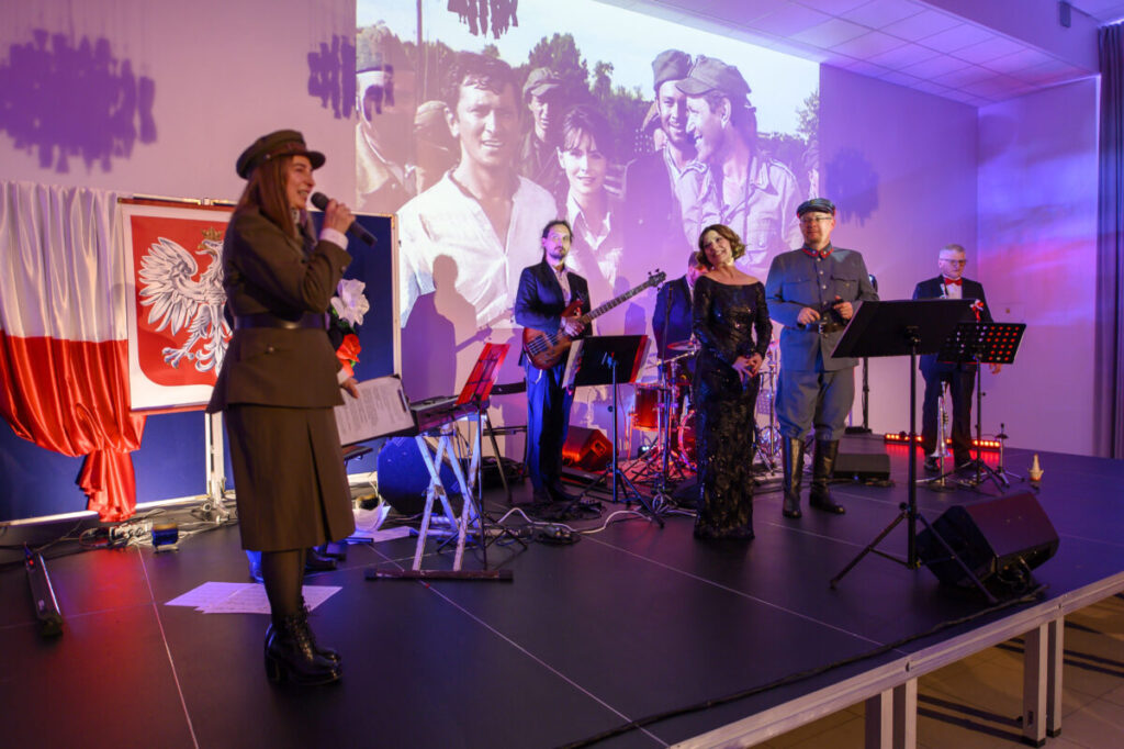 Na scenie zespoły w eleganckich strojach wojskowych oraz cywilnych, z instrumentami. Tło zawiera biało-czerwoną flagę i projekcję archiwalnych zdjęć. Oświetlenie w ciepłych odcieniach fioletu.