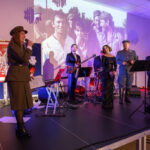 Na scenie zespoły w eleganckich strojach wojskowych oraz cywilnych, z instrumentami. Tło zawiera biało-czerwoną flagę i projekcję archiwalnych zdjęć. Oświetlenie w ciepłych odcieniach fioletu.