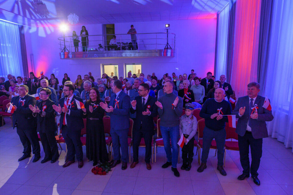 Sala wypełniona ludźmi świętującymi, trzymającymi polskie flagi. Dominują kolory czerwony, biały i niebieski. W tle widoczny balkon z osobami filmującymi.