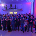 Sala wypełniona ludźmi świętującymi, trzymającymi polskie flagi. Dominują kolory czerwony, biały i niebieski. W tle widoczny balkon z osobami filmującymi.