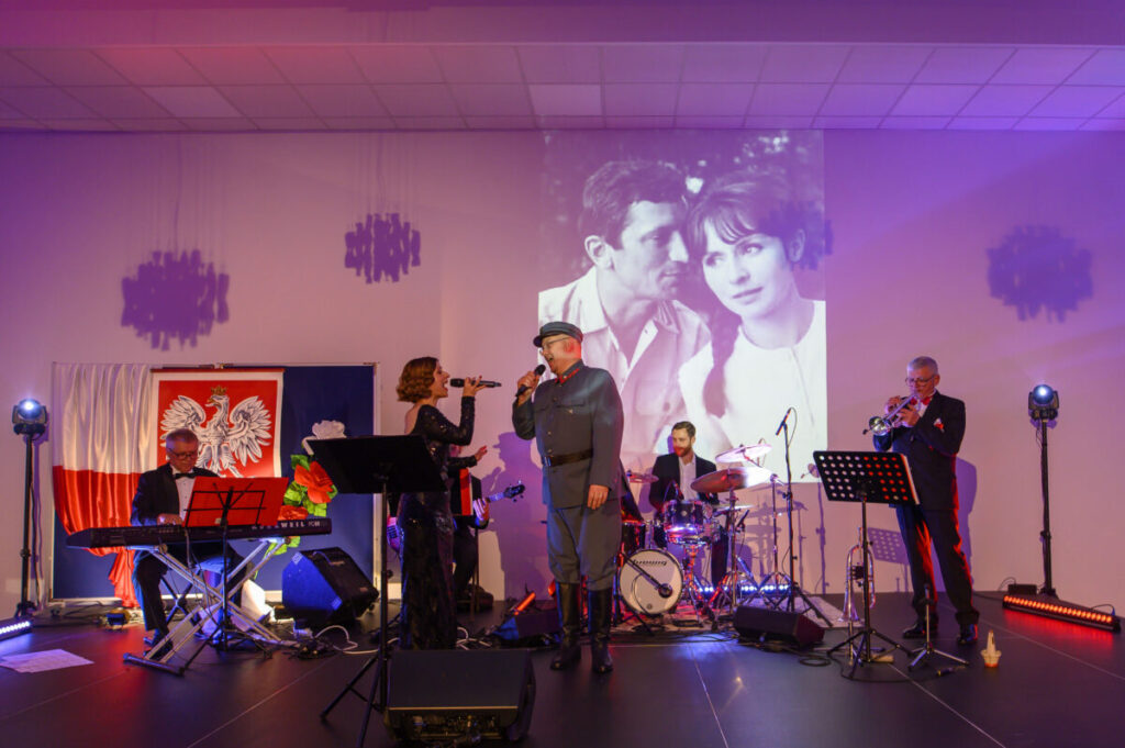 Na scenie zespół muzyczny z instrumentami, wokalistą w mundurze. Tło: biało-czerwona flaga, czarno-białe zdjęcie pary. Kolorowe światła tworzą atmosferę koncertu.