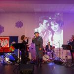 Na scenie zespół muzyczny z instrumentami, wokalistą w mundurze. Tło: biało-czerwona flaga, czarno-białe zdjęcie pary. Kolorowe światła tworzą atmosferę koncertu.