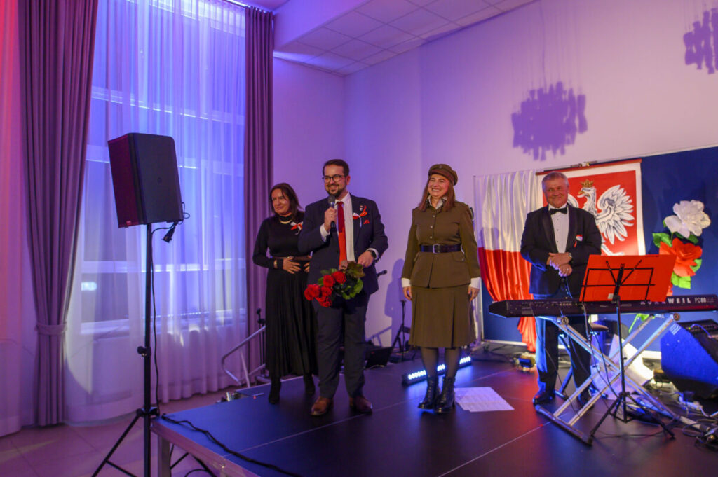 Grupa ludzi stoi na scenie, jeden z nich trzyma mikrofon. W tle są biało-czerwone dekoracje. Scena jest oświetlona fioletowym światłem.