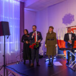 Grupa ludzi stoi na scenie, jeden z nich trzyma mikrofon. W tle są biało-czerwone dekoracje. Scena jest oświetlona fioletowym światłem.