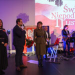 Na zdjęciu grupa osób na scenie, przy mikrofonie, z flagą Polski w tle. Dominują kolory czerwony, biały i granatowy. W tle widnieje napis Święto Niepodległości, 11 Listopada.