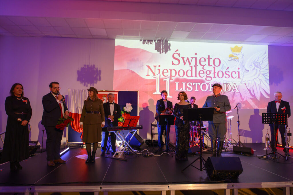 Grupa ludzi na scenie w eleganckich strojach. W tle białe płótno z napisem Święto Niepodległości 11 Listopada. Oświetlenie w tonacji fioletowej.