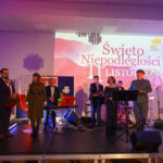 Grupa ludzi na scenie w eleganckich strojach. W tle białe płótno z napisem Święto Niepodległości 11 Listopada. Oświetlenie w tonacji fioletowej.