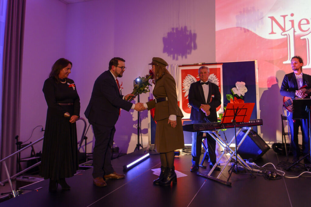 Na scenie mężczyzna wręcza kobiecie kwiaty; w tle flaga z orłem. Dominuje fioletowe oświetlenie. Obok stoją muzycy z instrumentami.