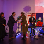 Na scenie mężczyzna wręcza kobiecie kwiaty; w tle flaga z orłem. Dominuje fioletowe oświetlenie. Obok stoją muzycy z instrumentami.