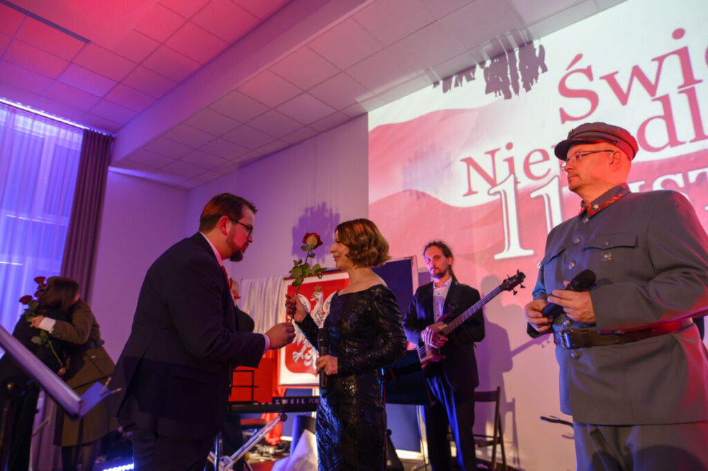 Na scenie widać ludzi w eleganckich strojach, trzymających czerwone róże. Tło jest jasne z napisem „Święto Niepodległości 11 listopada”. Panuje uroczysta atmosfera z kolorowym oświetleniem.