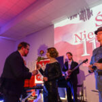 Na scenie widać ludzi w eleganckich strojach, trzymających czerwone róże. Tło jest jasne z napisem „Święto Niepodległości 11 listopada”. Panuje uroczysta atmosfera z kolorowym oświetleniem.