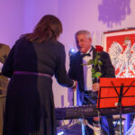 Mężczyzna w smokingu trzyma różę i podaje rękę kobiecie na tle godła Polski. Kolory dominujące: czerń i czerwony. W tle jasne ściany.