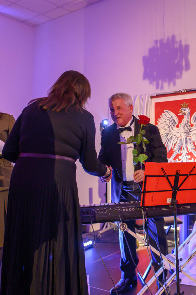 Mężczyzna w smokingu trzyma różę i podaje rękę kobiecie na tle godła Polski. Kolory dominujące: czerń i czerwony. W tle jasne ściany.