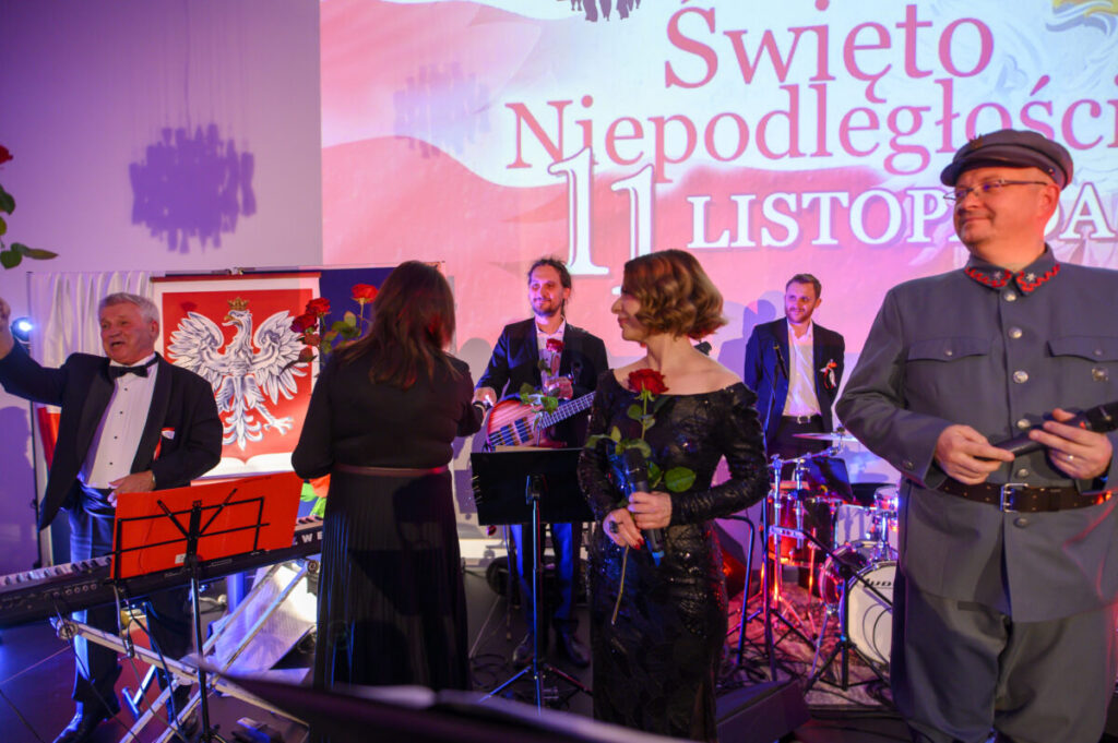 Grupa ludzi świętuje, trzymając róże. Tło jest jasne z widocznym polskim orłem i napisem Święto Niepodległości. Scena oświetlona w ciepłych barwach.