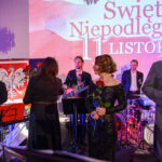 Grupa ludzi świętuje, trzymając róże. Tło jest jasne z widocznym polskim orłem i napisem Święto Niepodległości. Scena oświetlona w ciepłych barwach.