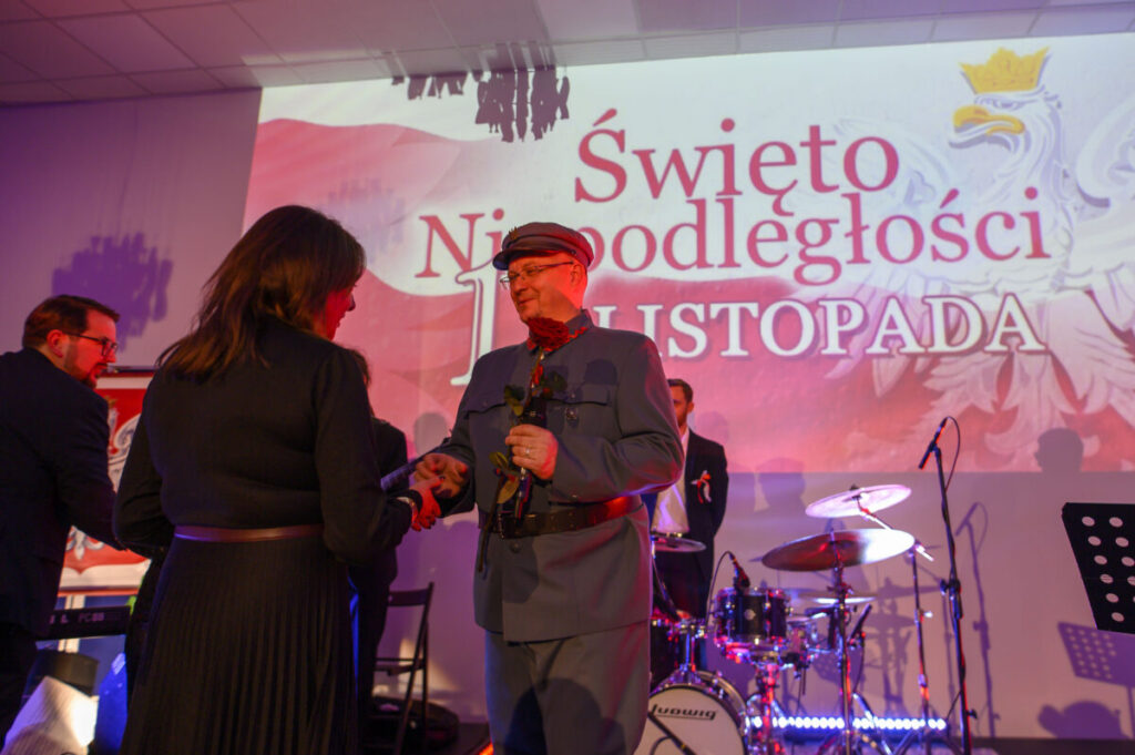 Na zdjęciu widać wydarzenie z okazji Święta Niepodległości. Osoba w mundurze wręcza różę, w tle bęben i napis. Kolory dominujące to czerwony i biel.