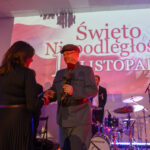 Na zdjęciu widać wydarzenie z okazji Święta Niepodległości. Osoba w mundurze wręcza różę, w tle bęben i napis. Kolory dominujące to czerwony i biel.