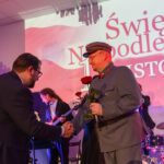 Mężczyźni w garniturach i mundurze podają sobie dłonie na tle sceny. Dominują kolory czerwony, szary, niebieski. W tle napis Święto Niepodległości.