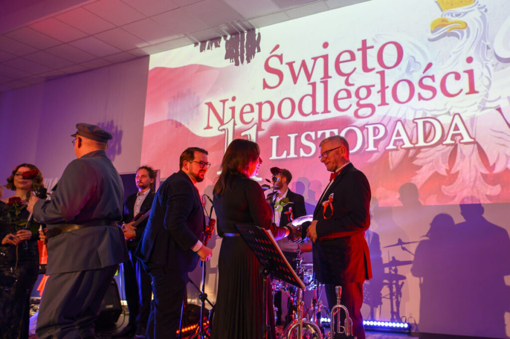Grupa ludzi na scenie podczas uroczystości z okazji Święta Niepodległości. Tło w biało-czerwonych barwach z dużym napisem. Oświetlenie sceniczne tworzy ciepłą atmosferę.
