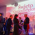 Grupa ludzi na scenie podczas uroczystości z okazji Święta Niepodległości. Tło w biało-czerwonych barwach z dużym napisem. Oświetlenie sceniczne tworzy ciepłą atmosferę.