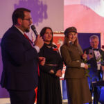 Grupa ludzi na scenie, kobieta w mundurze trzyma różę. Tło z flagą Polski, ciepłe oświetlenie. Mężczyzna z mikrofonem.