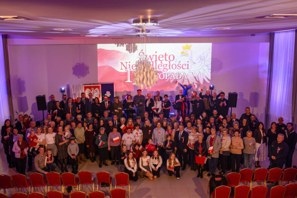 Duża grupa ludzi w sali z flagami Polski, świętująca 11 listopada. Dominują biało-czerwone kolory. W tle widoczny baner z napisem „Święto Niepodległości”.