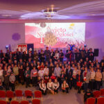 Duża grupa ludzi w sali z flagami Polski, świętująca 11 listopada. Dominują biało-czerwone kolory. W tle widoczny baner z napisem „Święto Niepodległości”.