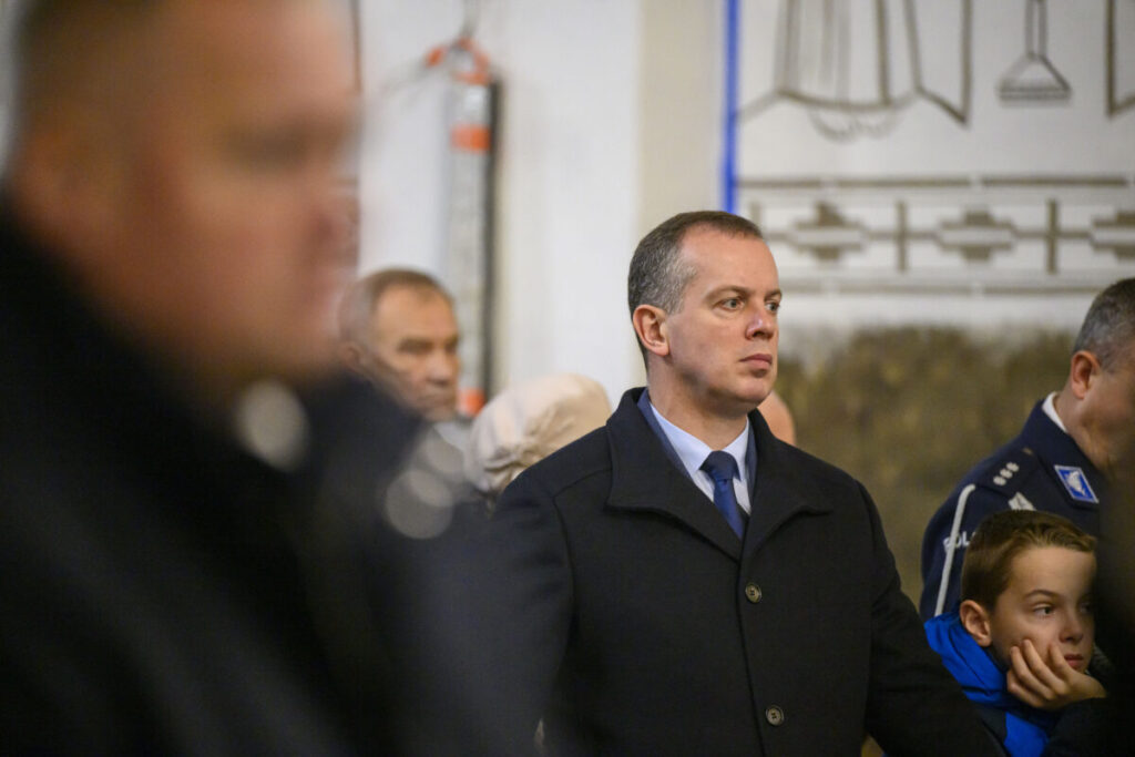 Na zdjęciu widoczny jest mężczyzna w ciemnym płaszczu. W tle inne osoby oraz rozmyte dekoracje na ścianie. Zdjęcie jest kolorowe.