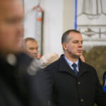 Na zdjęciu widoczny jest mężczyzna w ciemnym płaszczu. W tle inne osoby oraz rozmyte dekoracje na ścianie. Zdjęcie jest kolorowe.