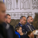 Na zdjęciu jest grupa ludzi w mundurach i dzieci, w tle ściana z rysunkami. Przeważają kolory niebieski i szary. Scena ma oficjalny charakter.