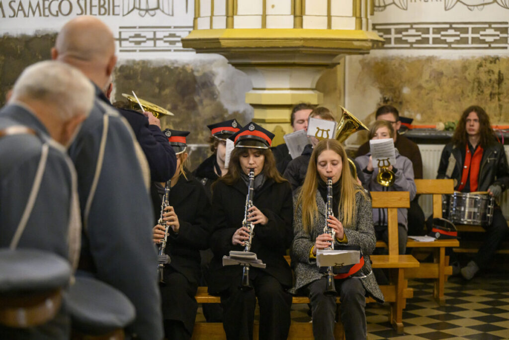 Muzycy orkiestry dętej grają w kościele. Ubrani w granatowe mundury z czerwonymi akcentami na tle ozdobnych ścian. Klarneciści siedzą z przodu, inni z tyłu mają instrumenty dęte.