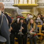 Muzycy orkiestry dętej grają w kościele. Ubrani w granatowe mundury z czerwonymi akcentami na tle ozdobnych ścian. Klarneciści siedzą z przodu, inni z tyłu mają instrumenty dęte.