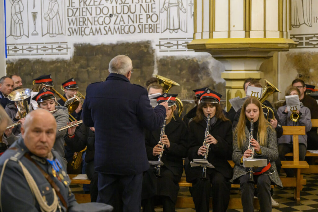 Orkiestra dęta w kościele, muzycy w ciemnych uniformach i czerwonych czapkach. Dyrygent w ciemnym płaszczu na środku. Ściany jasne, zdobione napisami i ornamentami.