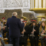 Orkiestra dęta w kościele, muzycy w ciemnych uniformach i czerwonych czapkach. Dyrygent w ciemnym płaszczu na środku. Ściany jasne, zdobione napisami i ornamentami.